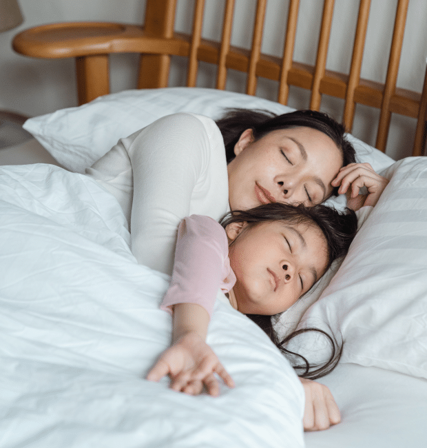 A mother and her child are peacefully sleeping together in bed, wrapped snugly in a white blanket. The child nestles comfortably in the mother's arms, both with their eyes gently closed. The soft hum of nearby labs creates a soothing background, enhancing the tranquility of their restful sleep.